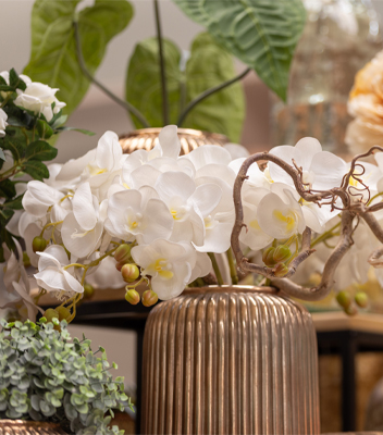 Eleganti orchidee bianche inserite in un vaso di design dal colore oro. A completare la composizione dei rami sinuosi senza foglie.