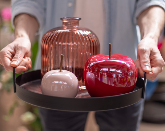 OggettisticaMele+Vaso Due mani sorreggono un vassoio in metallo nero su cui sono poggiati tre oggetti. Nello specifico si vedono due mele decorative, una piccola color rosa e una più grande rossa, ed un vaso di design color rosa
