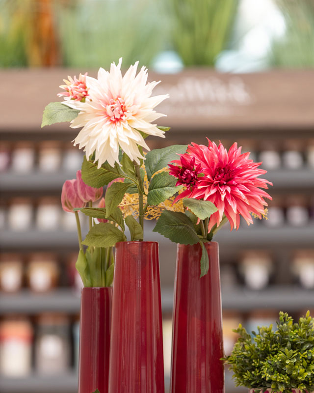 Dalie sintetiche nelle colorazioni rosa pallido e rosso inserite in vasi di design lunghi di colore bordeaux. Sullo sfondo candele