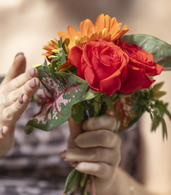 Primo piano della realizzazione di un bouquet, una mano è intenta a sorreggere i fiori mentre l'altra li posiziona e li sistema delicatamente. Nel bouquet sono state inserite 2 rose rosse, qualche gerbera arancione delle foglie.