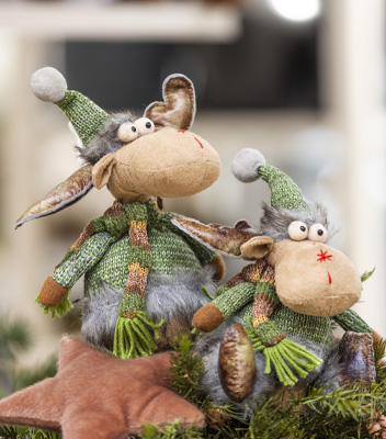 Medium shot di due renne di peluches color grigio. Le due renne poste una accanto all'altra indossa uno un cappellino di lana verde, un maglioncino, sempre in lana, verde e una sciarpa.