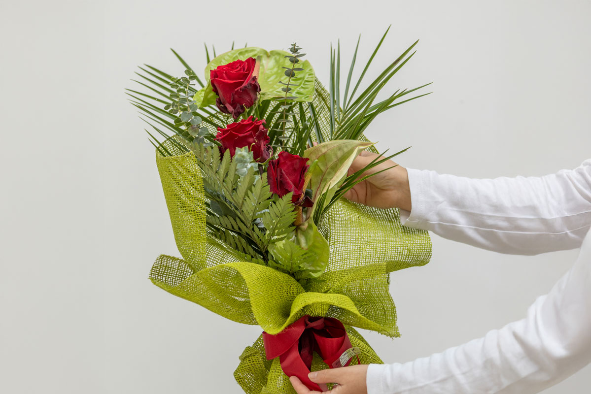 Al centro un bouquet composto da 3 rose rosse, 2 anturio verdi, felci ed eucalipto. Il bouquet è stato impreziosito da una fiocco rosso e una carta verde, in abbinamento ai fiori di anturio. Sulla destra si vedono le mani di una persona intente a sistemare leggermente il fiocco e un fiore