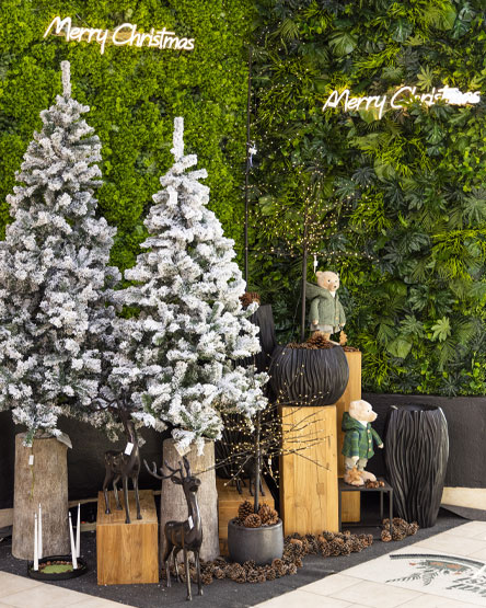 Allestimento natalizio con abeti innevati, decorazioni luminose 'Merry Christmas', orsetti decorativi, vasi moderni e dettagli di pigne su uno sfondo di parete verde.