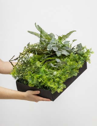 pareteVerde_artificiale Mani che reggono un modulo di parete verde artificiale con piante realistiche, ideale per decorazioni interne ed esterne.