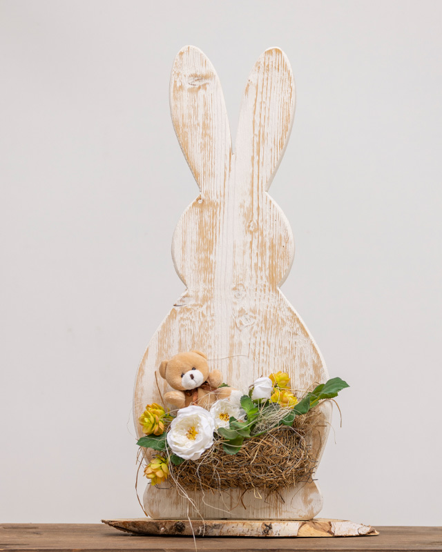 Decorazione pasquale in legno a forma di coniglio con fiori bianchi e gialli in un nido di paglia.