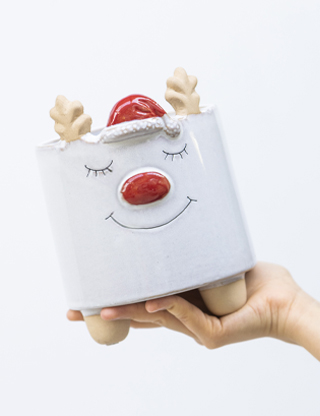 Vivai-Tomasi-vaso-natalizio Vaso decorativo bianco a forma di renna che sorride. La renna ha un grosso naso rosso e indossa un cappellino natalizio come quello di Babbo Natale.